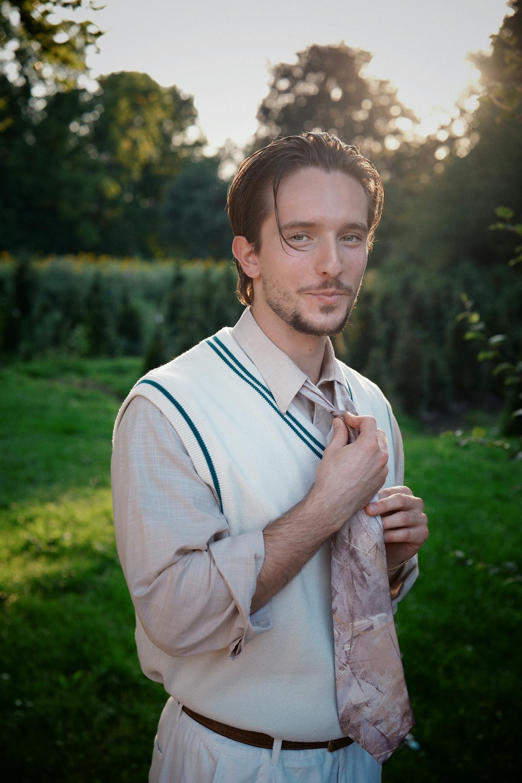 A young man adjusts his tie in a serene garden setting during sunset, exuding elegance and style.